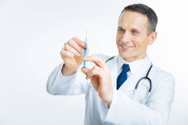 Radiant mature doctor looking at syringe - Фото, изображение