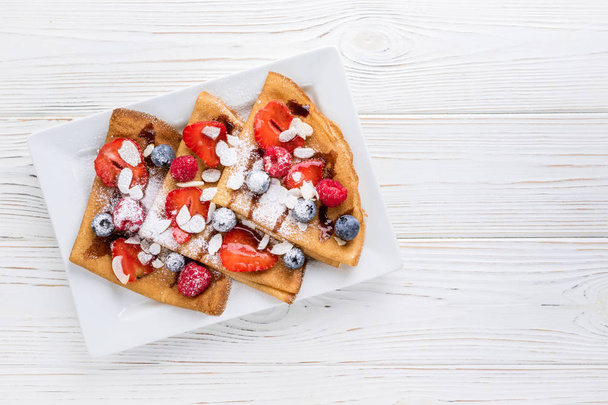 Crepes with strawberries, blueberries, almond and chocolate sauce on white wooden background. Top view - Photo, Image