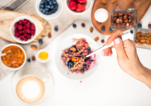 Widok z góry wyświetlone ręce jeść owsiankę z miód orzechy, jagody na biały drewniany stół selektywne skupić, niewyraźne tło dzień dobry - tło zdrowy śniadanie - Zdjęcie, obraz