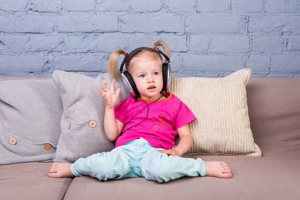 Дитяча дівчинка сидить на дивані з подушками і слухає музику у великих навушниках, одягнених на голову
. - Фото, зображення