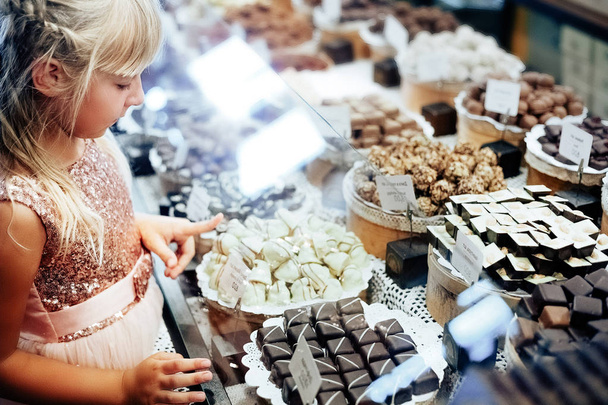 Uma menina loira está parada perto de uma vitrine com uma variedade de doces. A criança escolhe doces
 - Foto, Imagem