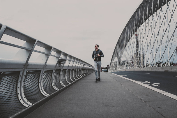 Young man running on the modern bridge in the city, listening music on smartphone - 写真・画像