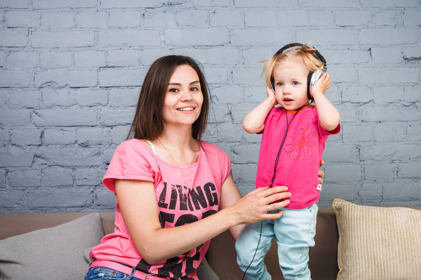 Mom and daughter listen to music in big headphones put on their head, sitting on the sofa. Holds the phone. - Photo, Image