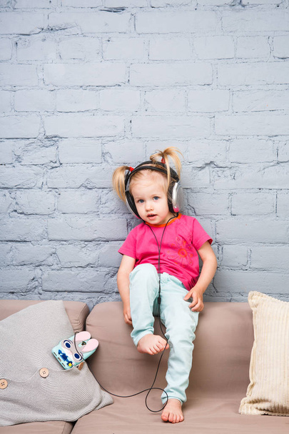 ein kleines lustiges Mädchen von zwei Jahren sitzt auf der Couch und hört Musik über Kopfhörer auf ihrem Kopf. hält ein Smartphone in der Hand - Foto, Bild