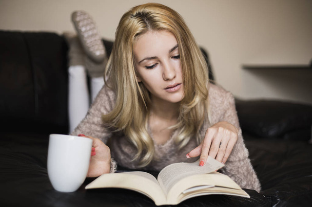 Nuori nainen kotona istuu ikkunan vieressä rentoutuen olohuoneessaan lukemassa kirjaa ja juomassa kahvia tai teetä - Valokuva, kuva