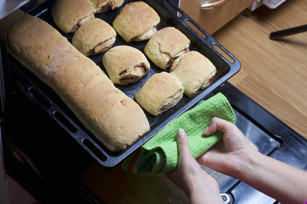 Preparation of cinnamon rolls. The woman takes a baking sheet out of the oven. There are ready buns and a roll with cinnamon on it. - Foto, immagini