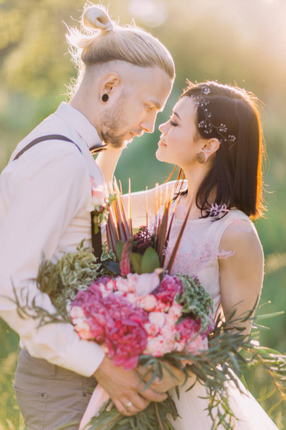The lovely close-up portrait of the newlyweds close to each other and holding the pink bouquet in the sunny forest. - Fotografie, Obrázek