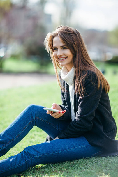 Beautiful smiling woman with phone sitting on a grass in nature - Фото, зображення