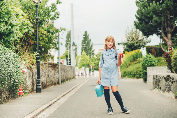 Outdoor portrait of funny little schoolgirl, wearing formal dress and backpack, holding lunch box and red apple. Back to school concept, film look toned image - Foto, immagini