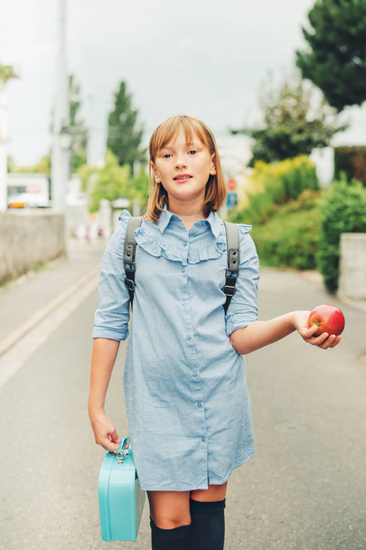 Outdoor portrait of funny little schoolgirl, wearing formal dress and backpack, holding lunch box and red apple. Back to school concept, film look toned image - Valokuva, kuva