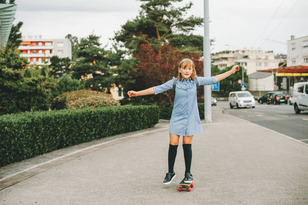 Funny little schoolgirl riding skateboard outside, back to school concept, film look toned image - Foto, afbeelding