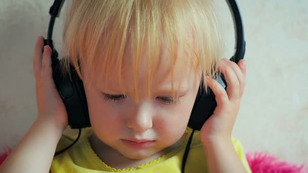 Крупним планом маленька смішна дівчинка два роки сидить на дивані, слухаючи музику на навушниках, одягнених на її голову
 - Кадри, відео