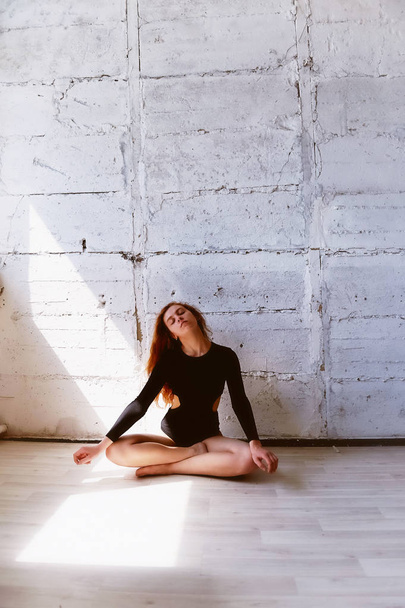 Model tests. Beautiful redhead girl with curly hair. Natural color. Demonstrates the flexibility for yoga, aerobics and ballet on the bright background of brick wall.Studio-loft. Dressed in a black - Fotó, kép
