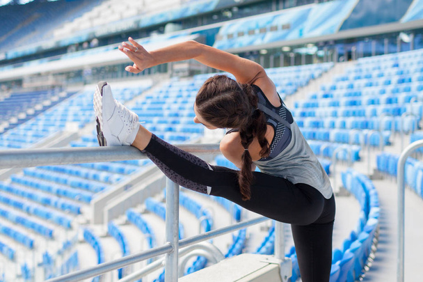 Young sporty girl with a tanned skin makes a banner in the stadium. The woman pulls the leg up, making the warm-up - Fotó, kép