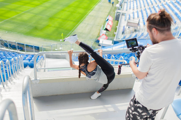 The cameraman shoots on a professional camera, as a sports girl kneads in a stadium against the background of a football field. A sexy woman holds on to a metal railing and raises her leg above head - Photo, Image
