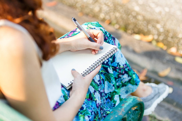 Cerca de la mano izquierda mujeres asiáticas con pluma escribir cuaderno y sentado en un banco de madera y un parque
. - Foto, imagen