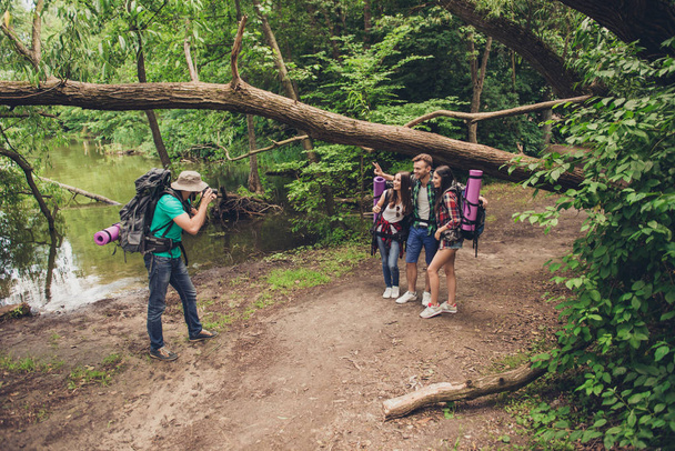 Male photographer is taking photo of his three friends near the lake in the spring wood, so beautiful nature! They are tourists, hiking jungle trail - Foto, imagen