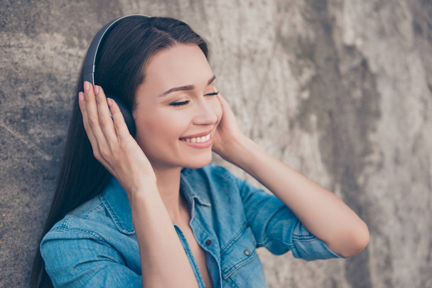 Charming young girl with toothy smile is so dreamy and happy, listening to her favourite song, holding the earphones, with closed eyes on concrete`s wall background - Photo, Image