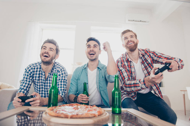 Yes! Team of winners! Bachelor men`s life. Low angle of three happy joyful men, sitting on sofa and playing video games with beer and pizza, smiling, gesturing, enjoying themselves - Photo, image