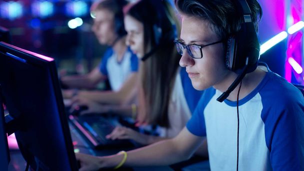 Professional Boy Gamer Plays in Video Game on a eSports Tournament/ Internet Cafe. He Wears Glasses and Headphones with Microphone. Other Girls and Boys Players Playing in Background. - Fotografie, Obrázek