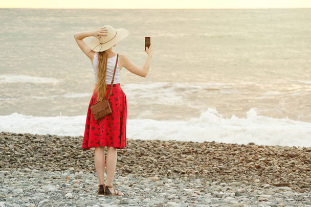 Meisje in de rode rok en hoed selfie op het strand op zee en hemel achtergrond maken. Uitzicht vanaf de achterkant - Foto, afbeelding