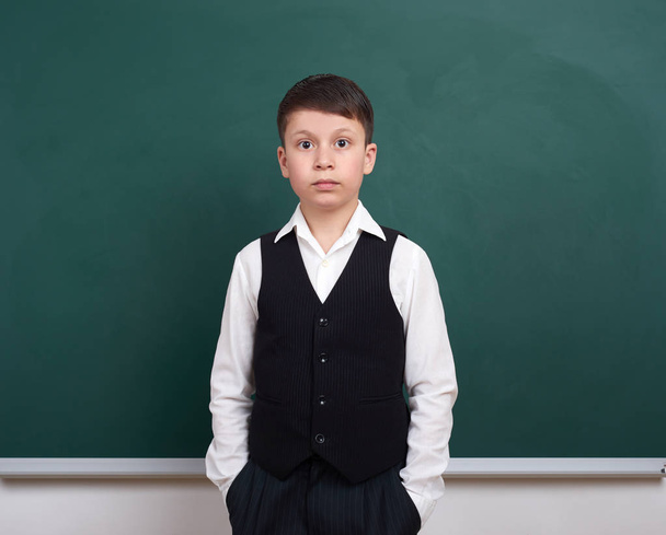 indifferent school boy portrait near green blank chalkboard background, dressed in classic suit, one pupil, education concept - Photo, Image