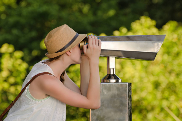 Girl in hat looks into big binoculars. Park in the background - Photo, Image