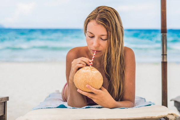 Young woman drinking coconut milk on Chaise-longue on beach. - Foto, afbeelding
