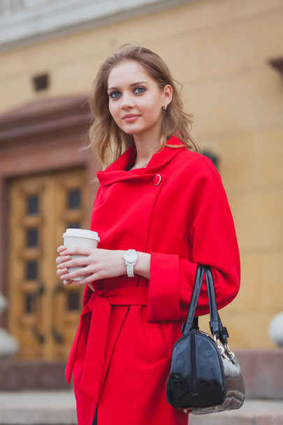 Portrait of a beautiful blonde girl with long hair in a red coat walking along the street. Girl holding coffee in hands - Foto, Imagen
