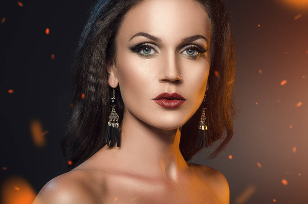 woman standing in the sparks of fire. Fashion portrait of brunette in black earrings on a dark background - Photo, Image