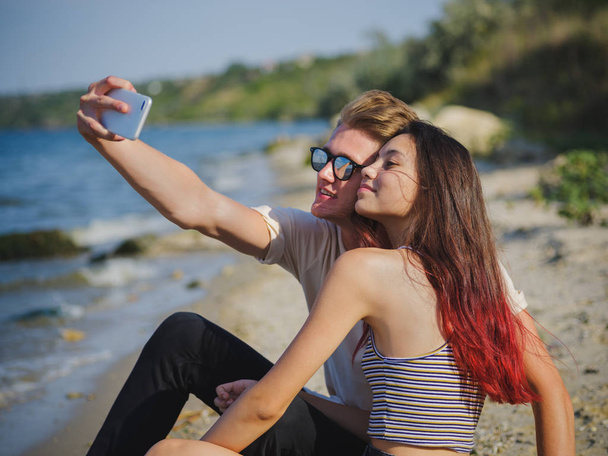 Close-up of a beautiful girl and a fellow taking a selfie on a riverbank on a natural blurred background. - Foto, Bild