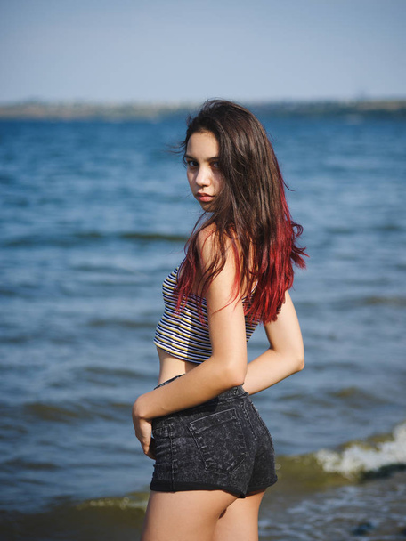 Confident young girl on a coast. Trendy, pretty girl model posing on a blurred water background. Teen fashion concept. - Foto, afbeelding