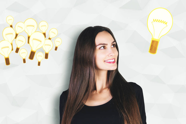 Attractive smiling young woman on polygonal wall background with drawn glowing light bulbs. Idea concept  - Foto, Imagen