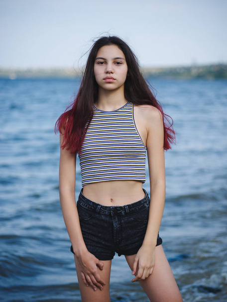Confident young girl on a coast. Trendy, pretty girl model posing on a blurred water background. Teen fashion concept. - Zdjęcie, obraz