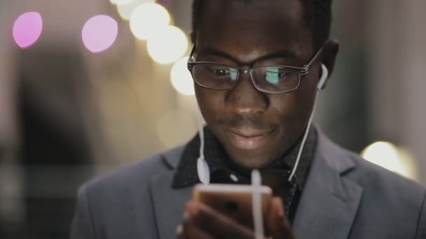 Young african student in classical suit listening to music with earphones outdoor - Metraje, vídeo