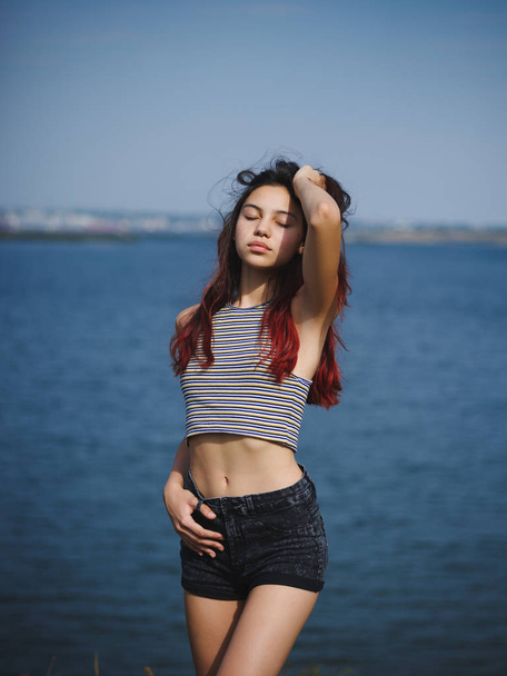 Sexy, cute young girl touching her hair. A teenager on a blurred river background. Beauty and style concept. Copy space. - Фото, изображение