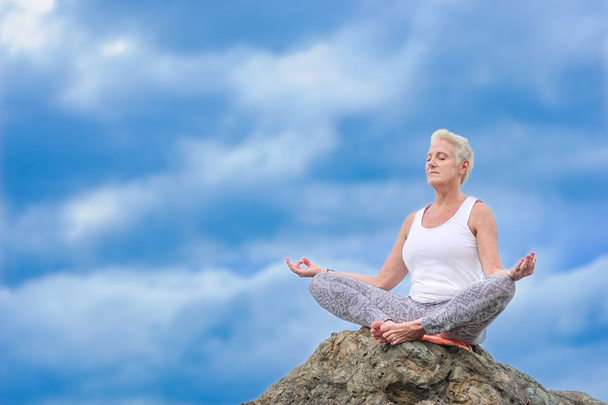 schöne ältere Frau sitzt oben auf einer Klippe und macht Yoga - Foto, Bild