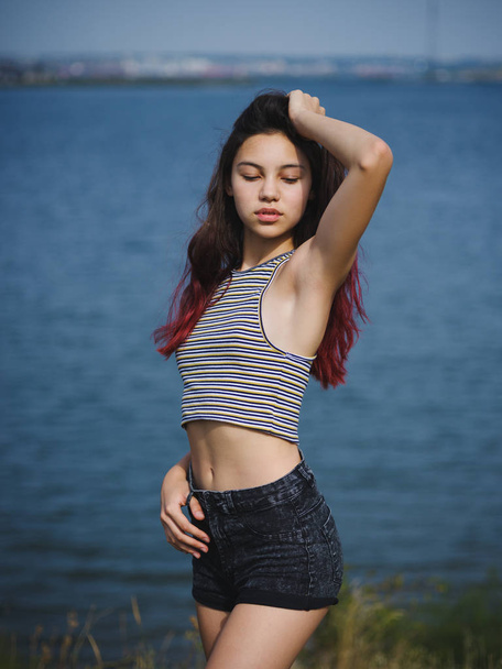 Sexy, cute young girl touching her hair. A teenager on a blurred river background. Beauty and style concept. Copy space. - Foto, afbeelding