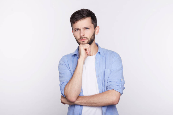 Young serious Caucasian man in shirt, holding hands on his chin, looking with thoughtful and skeptical expression on his face, suspecting of something, hesitating to make decision. Body language. - Photo, Image