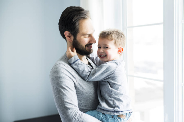 Happy young father holding cute boy standing near big window. - 写真・画像