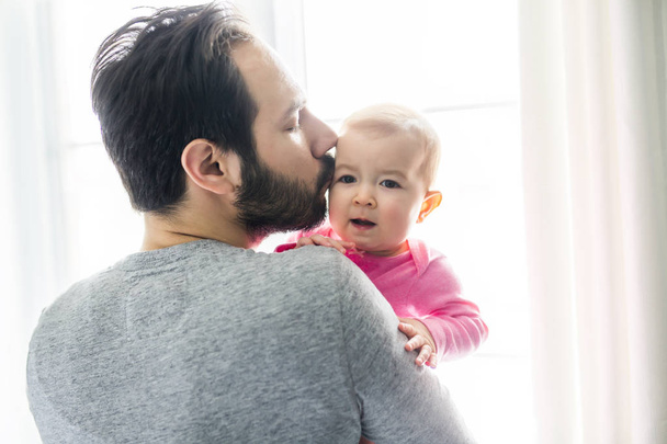 Happy young father holding cute baby girl standing near big window. - Photo, Image