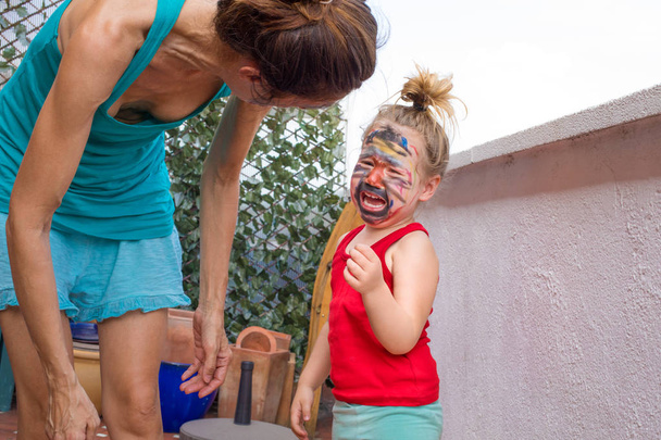 маленький ребенок с нарисованным лицом кричит рядом с матерью
 - Фото, изображение