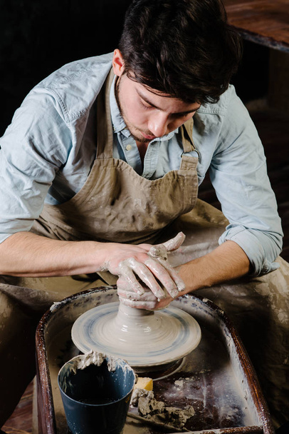 pottery, workshop, ceramics art concept - man working on potter's wheel with raw clay with hands, a male brunette sculpt a utensils near wooden table with tools, master in apron and a shirt - Photo, Image