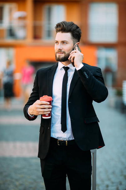 Confident young businessman using cell phone and drinking coffee in the city - Zdjęcie, obraz