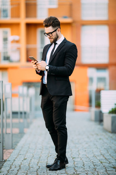 Hombre de negocios barbudo guapo en traje clásico está utilizando un teléfono inteligente mientras está de pie en el balcón de la oficina
 - Foto, imagen