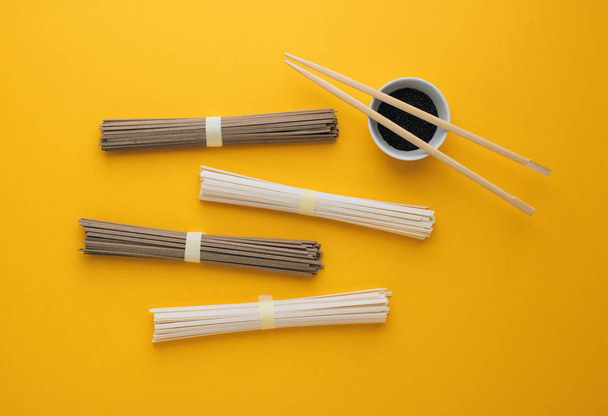 Asian noodles udon and soba, chopsticks, black sesame in a bowl on a bright yellow background. Minimalism. Flat lay.  - Photo, Image