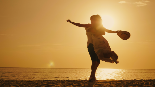男は、夕日と海の背景に彼の腕で女性を旋回です。一緒に幸せな休暇に到着 - 映像、動画