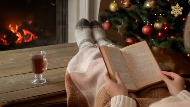 Closeup αργή κίνηση μήκος σε πόδηα του γυναίκα διαβάζοντας το βιβλίο δίπλα στο χριστουγεννιάτικο δέντρο και τζάκι - Πλάνα, βίντεο