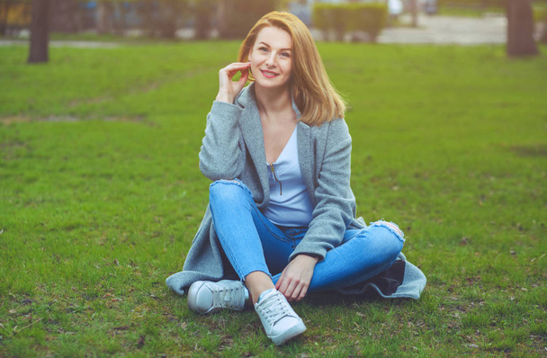 Приваблива, молода блондинка в сірому пальто і джинси сидять на траві
 - Фото, зображення