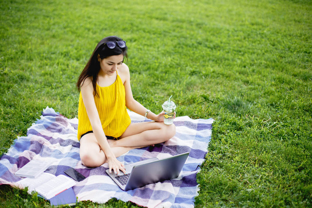 Beautiful young woman sitting on the green grass in park with her laptop. Student girl looking at the computer - Zdjęcie, obraz
